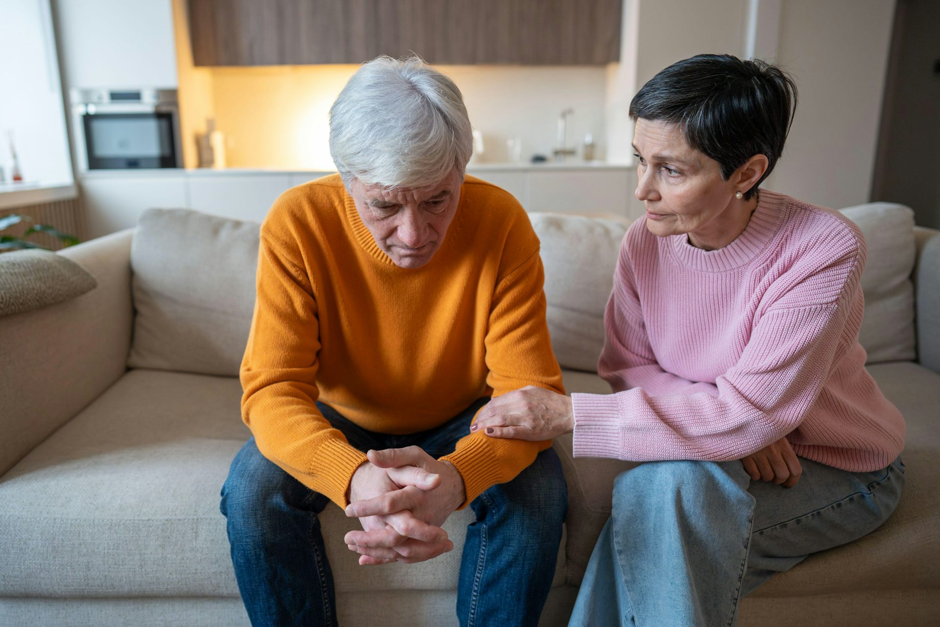 A husband and wife sit on a sofa. The wife looks concerned and grips the husband's forearm.