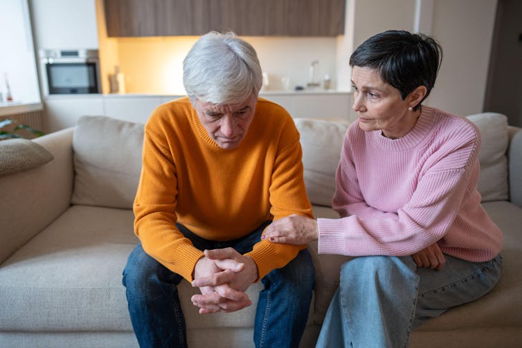 Um marido e uma esposa sentados em um sofá. A esposa parece preocupada e segura o antebraço do marido.