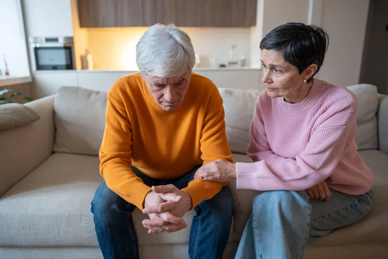 A husband and wife sit on a sofa. The wife looks concerned and grips the husband's forearm.