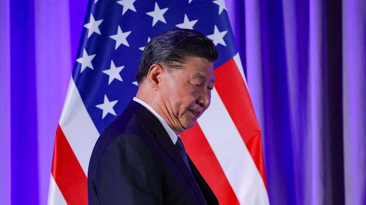 A man in a dark suit with a flag behind him.