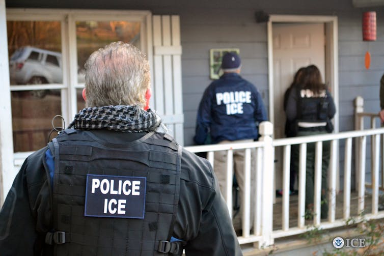 A man looks at a house and is seen from the back, wearing a black jacket that says 'Police ICE.' Two other people wearing similar jackets stand outside a door.