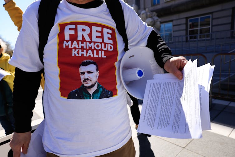 A person seen from the neck down wears a white shirt that says 'Free Mahmoud Khalil' and has a photo of a man on it.