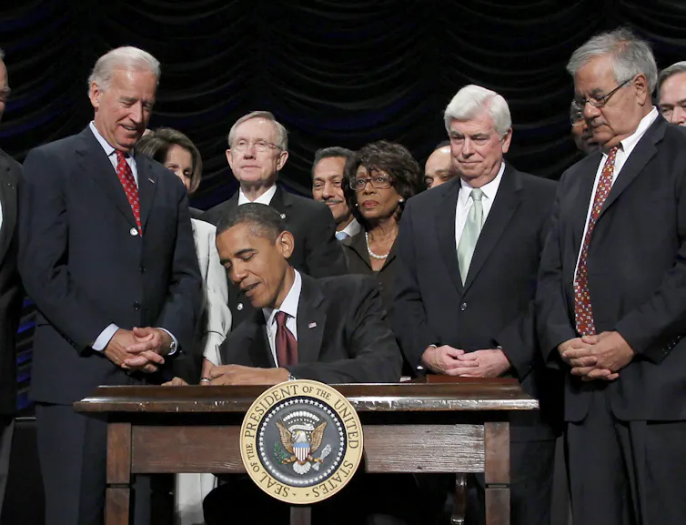 A Black man in a suit signs a document at a table with the U.S. presidential crest on it while a small crowd of people look on