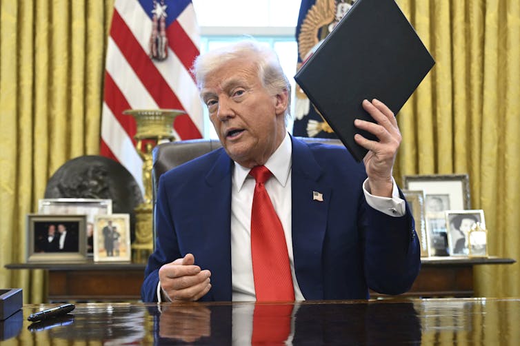 El hombre blanco con cabello blanco mantiene el folio cerrado mientras se sienta frente a la bandera de los Estados Unidos