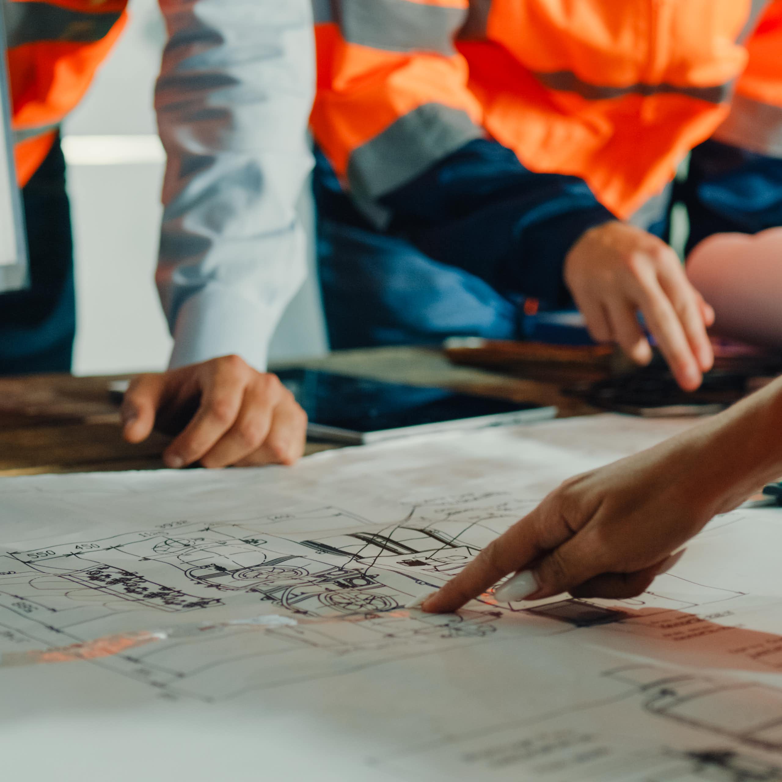 A group of people, one wearing a bright vest, examine and point to a blueprint diagram lying on a table.