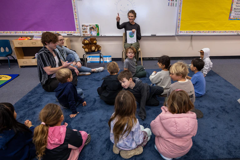 A teacher, holding a book, sits in a chair in a classroom in front of young children sitting on the ground in front of them. A smiling sun is drawn on the white board behind the teacher.