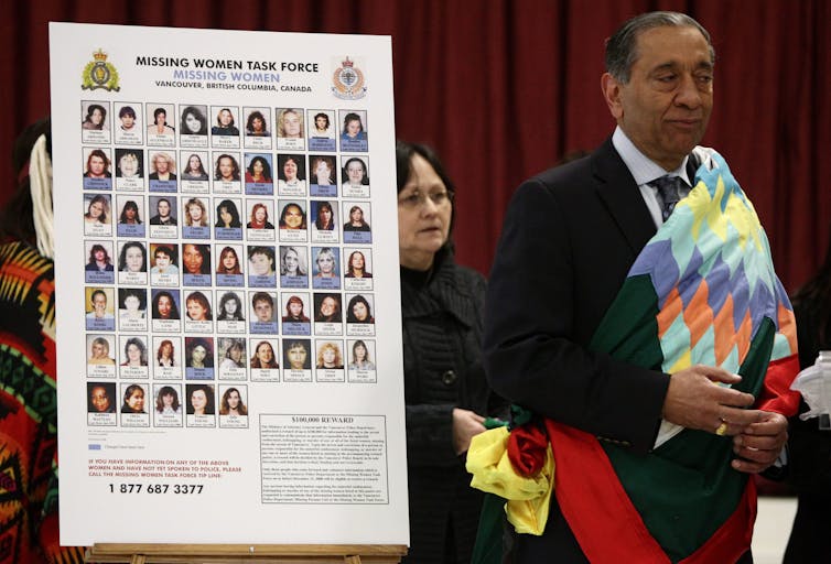 A older south asian man in a dark suit wrapped in a colouful Indigenous blanket stands next to a cut out with photos of women