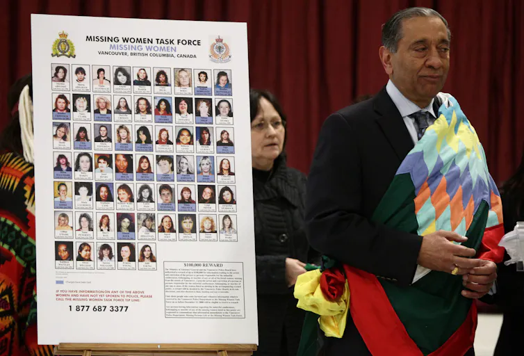 A older south asian man in a dark suit wrapped in a colouful Indigenous blanket stands next to a cut out with photos of women
