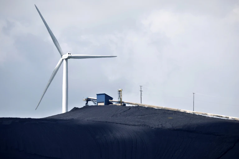 A large wind turbine behind a coal facility and coal pile.