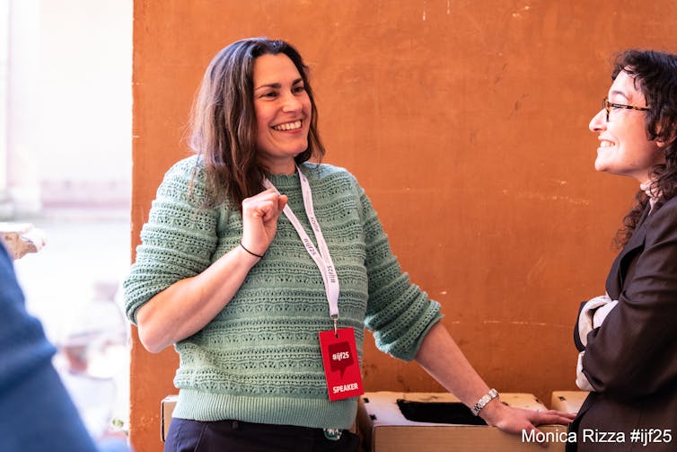 Anna woman in green jumper talks to another woman, standing in front of orange wall