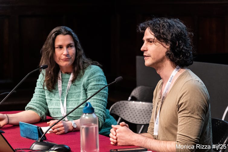 woman in green jumper Anna Turns sat at table interviewing Adam Levy with microphones