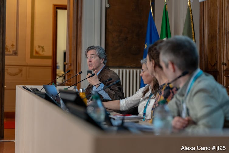 four people on panel, man on far left talking, flags and wood panelled walls in background