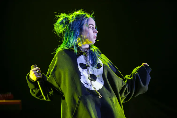 Teeanged girl wearing baggy black sweatshirt, holding a microphone, bathed in green light.