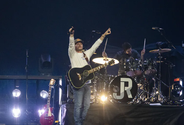 Young male guitar player wearing a hat and white shirt raises his arms in the air from stage in front of a drummer.