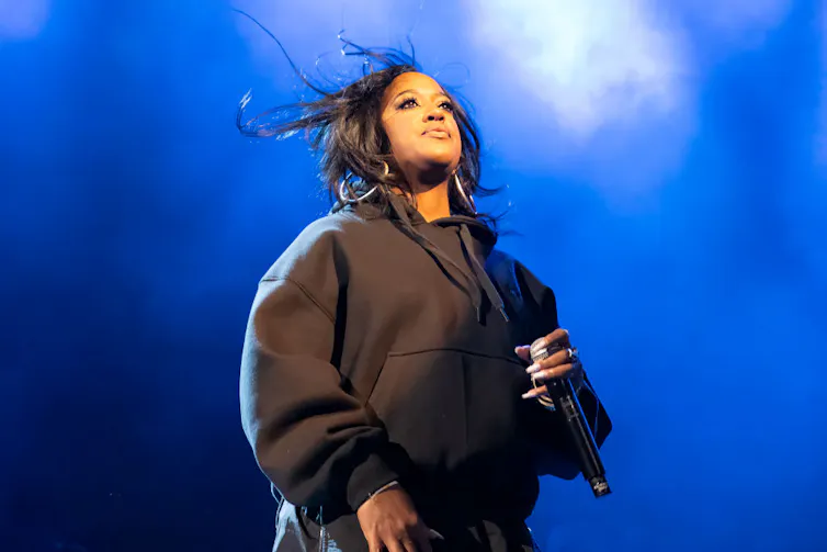 Woman wearing black sweatshirt holds a microphone on stage against a backdrop of blue lights.