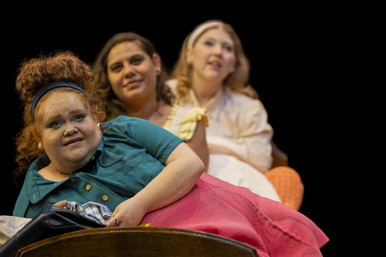 Production image: three young women sit on a bed.