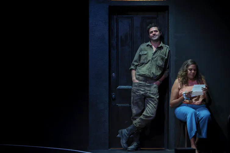 Production image: a man stands in a doorway; a woman sits and reads a letter.