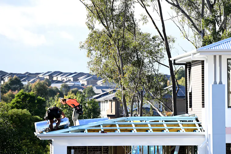 Two construction workers stand on the roof of a suburban house.