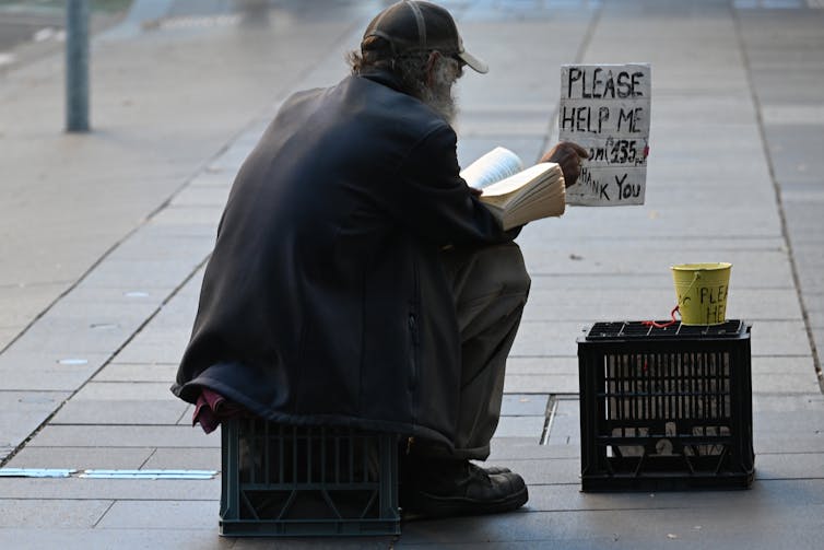 Dark clothed homeless man sitting on a footpath, holding a sign and reading a book
