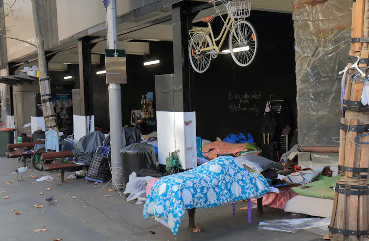 Cluttered footpath with a person sleeping under a blue and white blanket and beneath a bicycle hanging from a hook