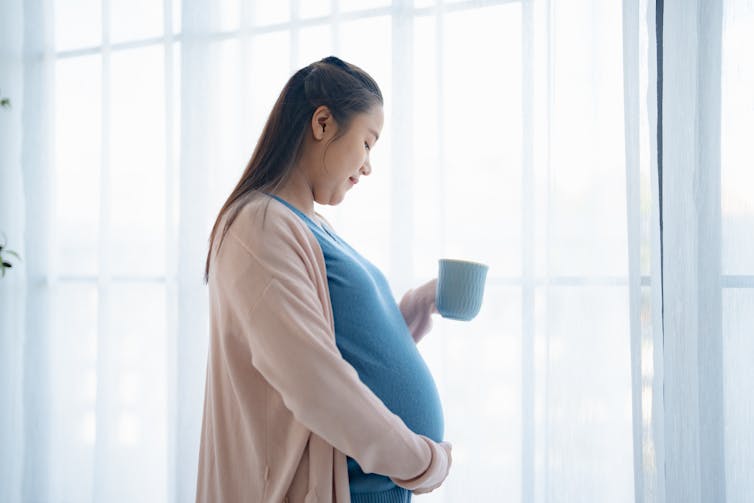 A pregnant woman wearing a tan cardigan and blue shirt holds her stomach with her right hand and mug in her left hand. She is next to a window.