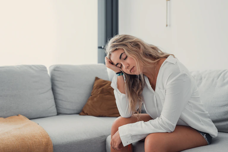 A tired blonde teenage girl sits on the edge of a couch with her head in her hand.