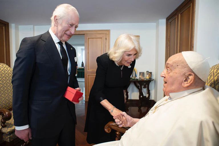The king and queen greet Pope Frances, who is seated in a wheelchair. Camilla is shaking the pope's hand, and the king is holding a small, red gift box.