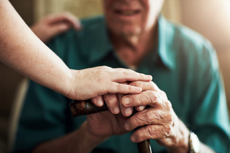 Close up of a carer's hands holding an elderly man's hands as he holds a cane