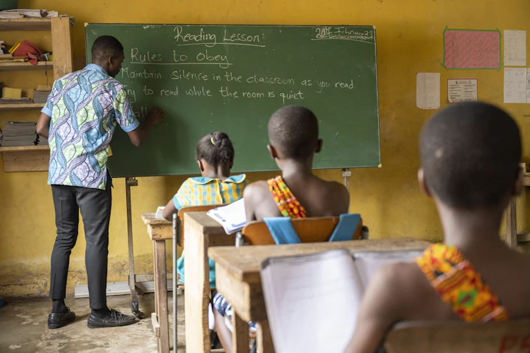 Man writing on a blackboard with children sitting at desks watching him