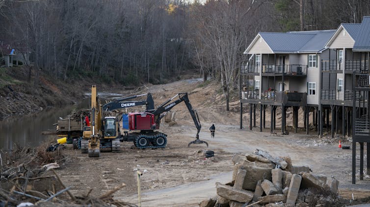 An apartment complex on stilts teeters at the edge of a river with visible scars from flooding.