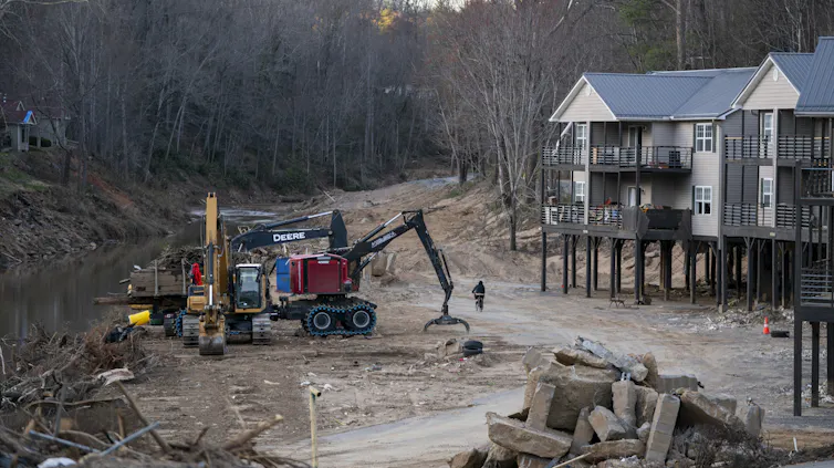 An apartment complex on stilts teeters at the edge of a river with visible scars from flooding.