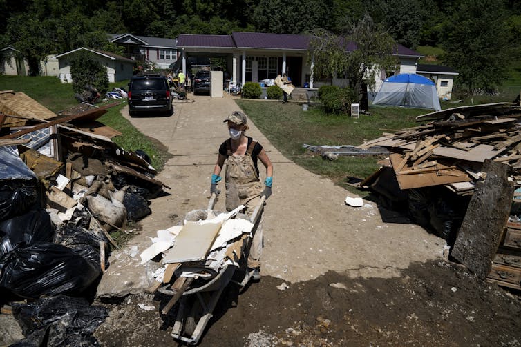 A woman wearing a mask walks a wheelbarrow full of debris out of a damaged home.