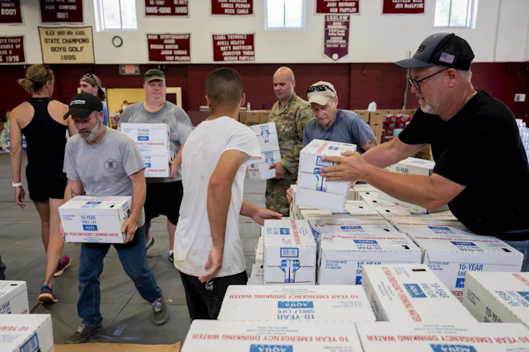 People lift boxes of water off a table in a school gymnasium; National Gardsmen are visible in the background.