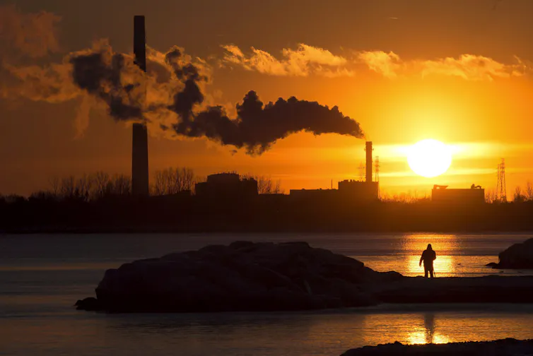 A man photographs a setting sun as smokestacks belch smoke into the orange sky