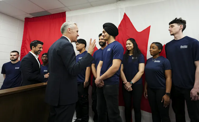 A white man in a dark suit talks to a young south asian man wearing a turban. Other young people stand in the background