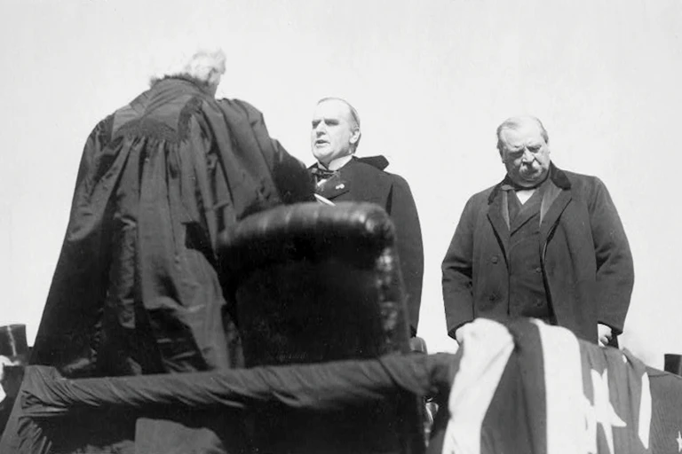 A man with his back to the camera and dressed in a black robe speaks to man facing him as another man looks away.
