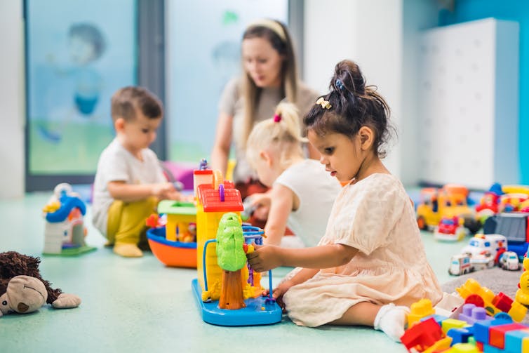 An early childhood educator sits on the floor with children as they play with toys.