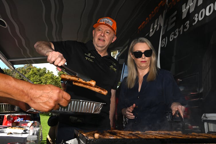 Anthony Albanese wears a cap and barbecues sausages next to his partner, Jodie.