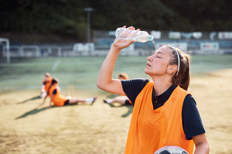 Young woman holding soccer ball pours water bottle on head.