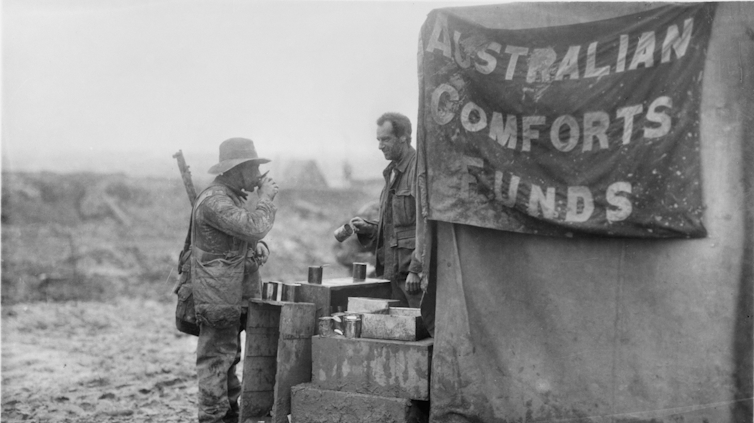 Black and white photograph: Two soldiers drink tea, a banner reads 'Australian Comforts Fund'