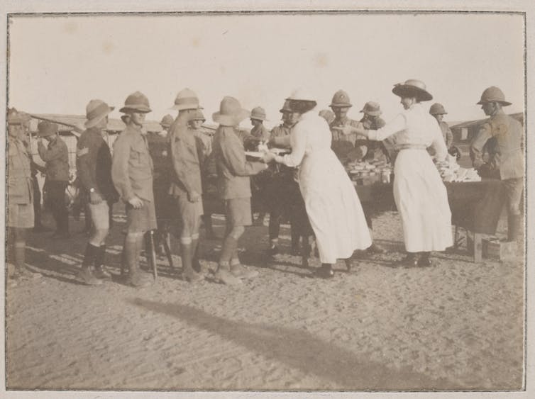 Sepia photograph: soldiers gathered around two women standing at a table handing out packages.