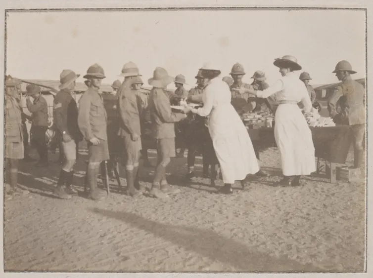 Sepia photograph: soldiers gathered around two women standing at a table handing out packages.