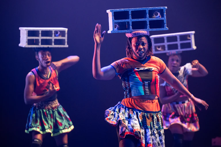 Three performers, men and women, dressed in skirts and balancing a large concrete brick on their heads like a hat.