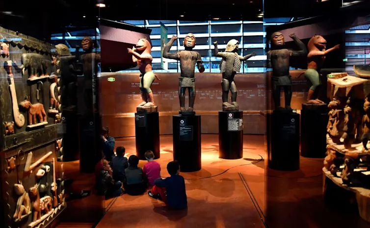 Children sit on the floor in a museum looking at three large statues of African warriors standing on plinths in pools of light.
