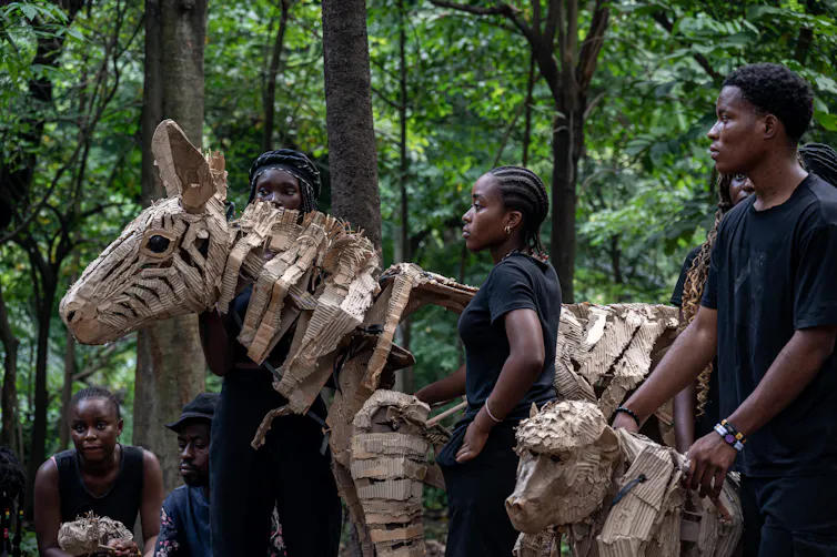 African men and women dressed in black stand in a forest holding life-size paper puppets of a zebra and a gorilla.