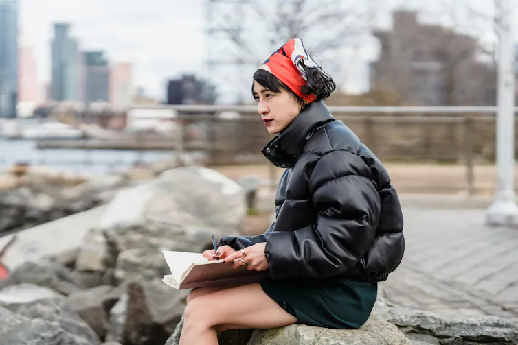 A girl sits on a low wall writing in a notebook.