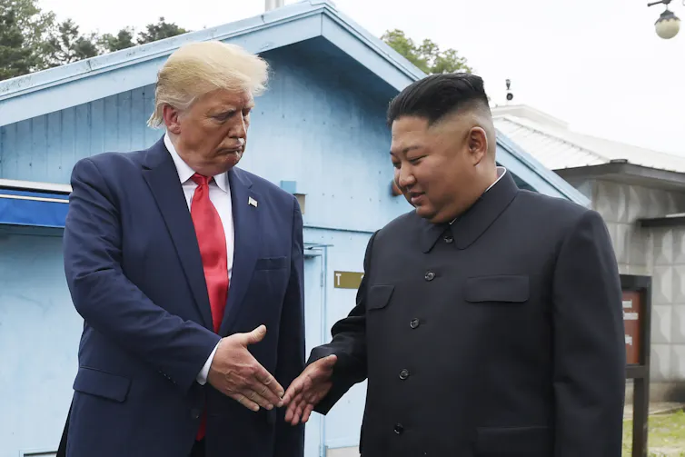 An older man with fluffy white-blong hair in a blue suit and red tie looks down as he's about to shake hands with a rotund Asian man.