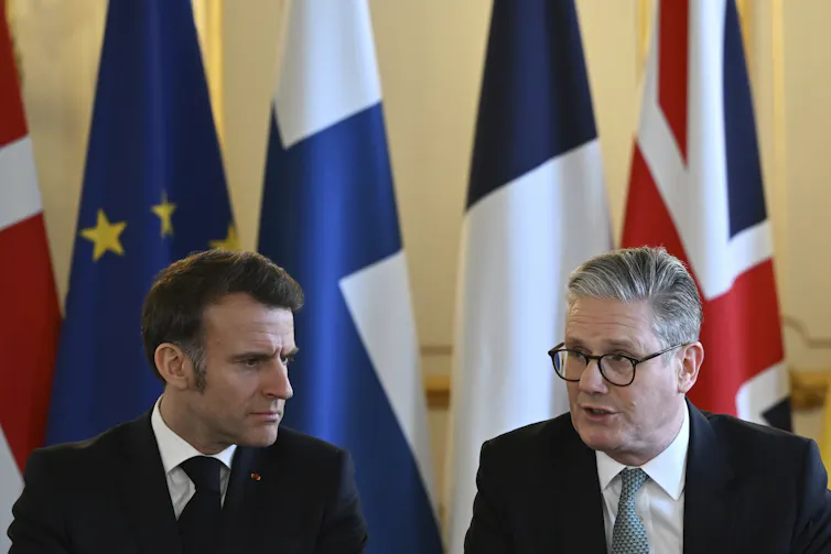 Two men, one with dark and one with grey hair and glasses, speak in front of a row of flags.