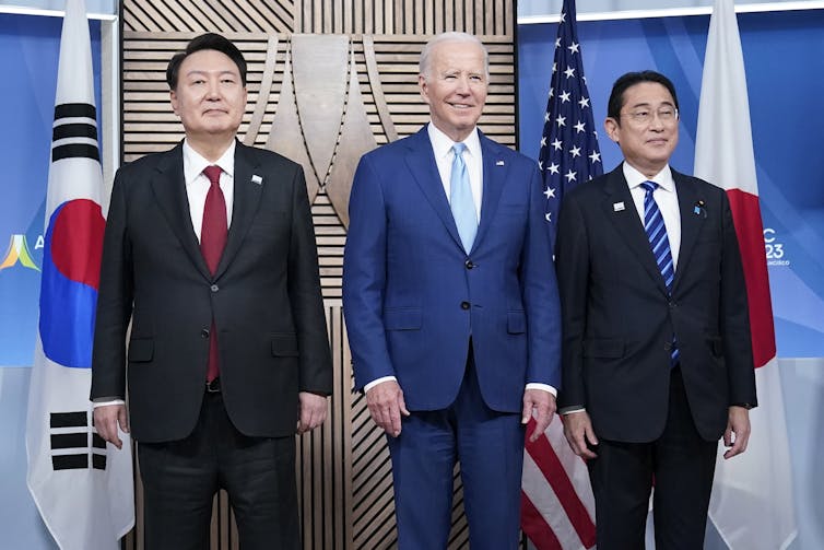 Dos hombres asiáticos de cabello oscuro y un hombre blanco de cabello gris mayor se paran en una serie que representa una fotografía.