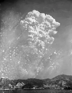 A black-and-white photo shows a massive mushroom cloud reaching high into the sky from a distance.